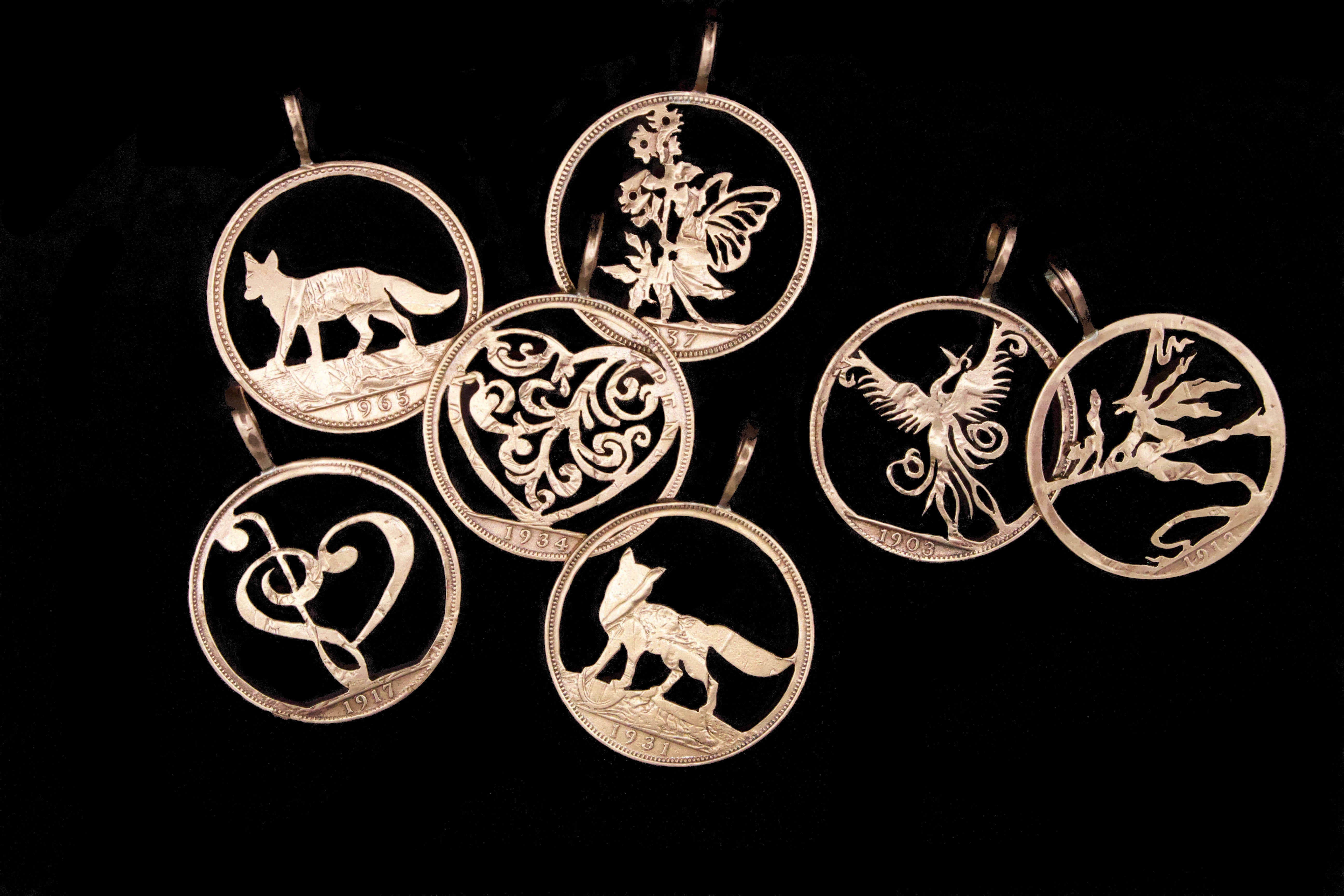 A collection of hand-cut old English penny pendants displayed against a black background, each intricately pierced with unique designs including animals, magical creatrures and hearts.