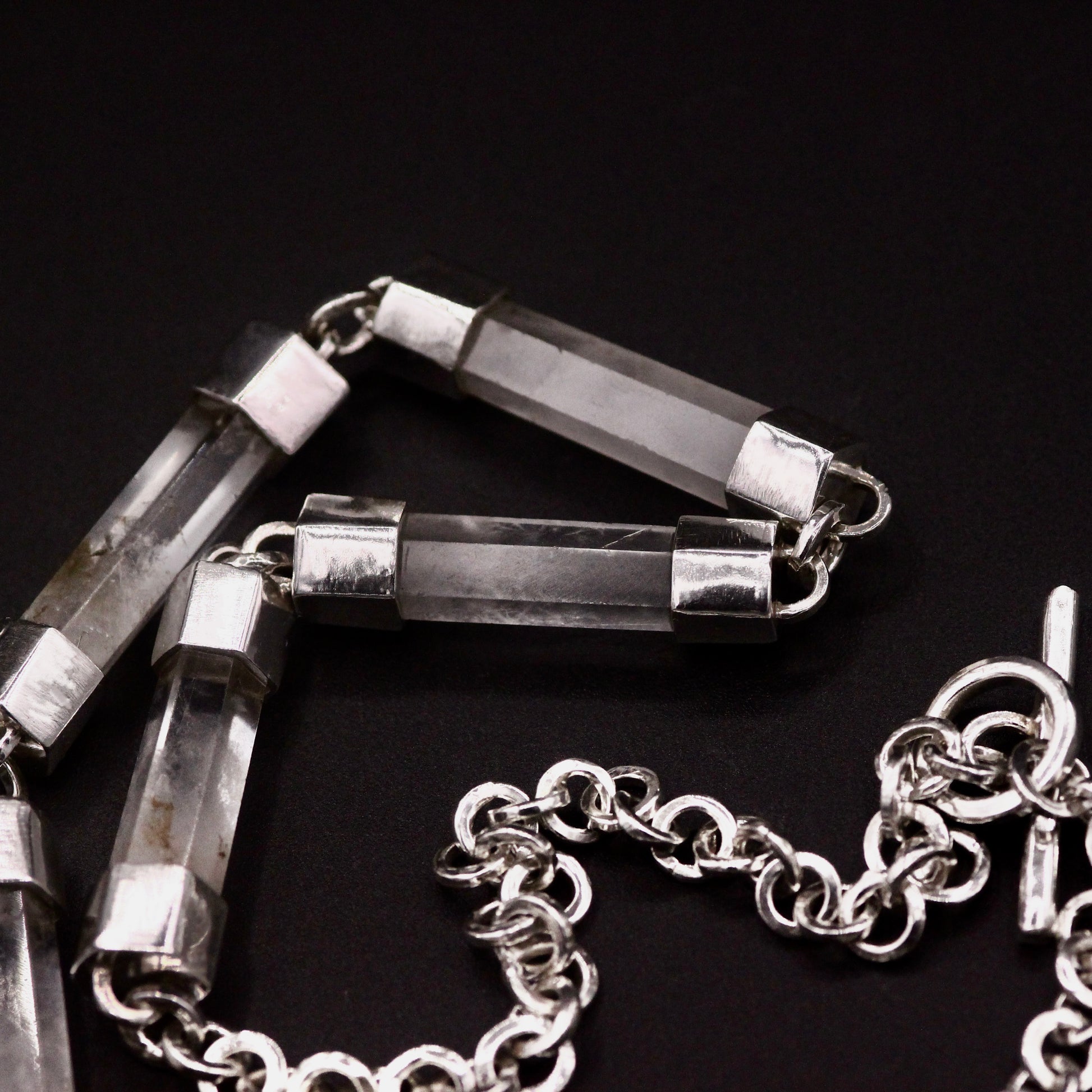 Handmade sterling silver and natural clear quartz crystal bar necklace coiled on black background, showing full length of crystal bar links transitioning into sterling silver belcher chain with T-bar toggle clasp, Thornhill Jewellery